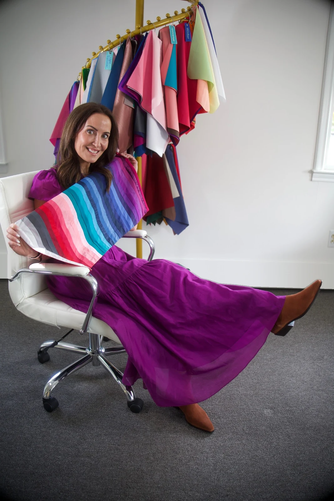 Kate in a magenta dress, seated in her studio and holding a fan of color-analysis drapes in front of a rack of seasonal swatches