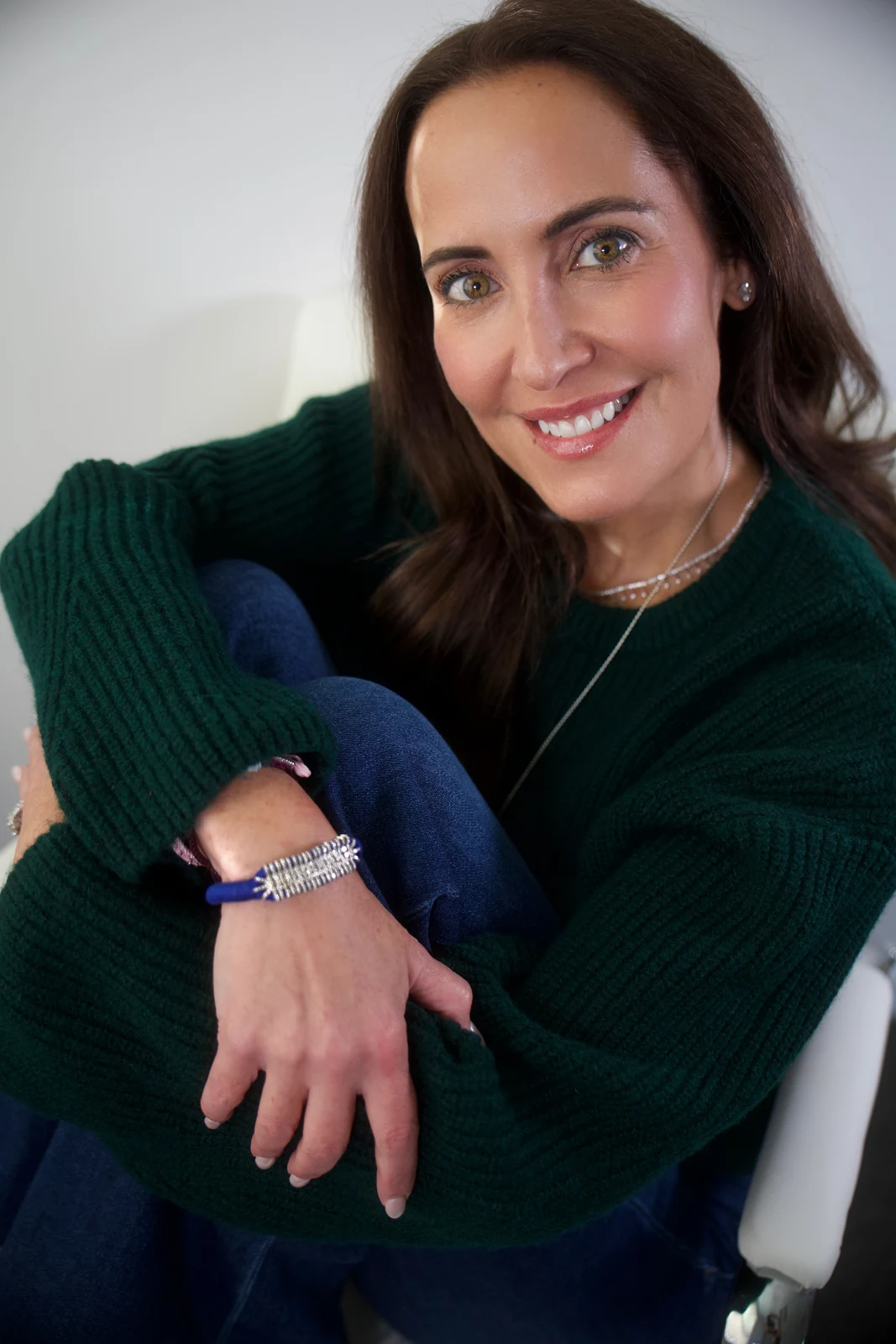 Kate smiling in a green knit sweater and jeans, arms wrapped around her knees, wearing her Royal Blue Slim Stacker diamond bracelet